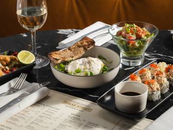 Bread Street Kitchen restaurant food spread featuring burrata, fresh salad, sushi rolls and white wine on black marble table with menu.