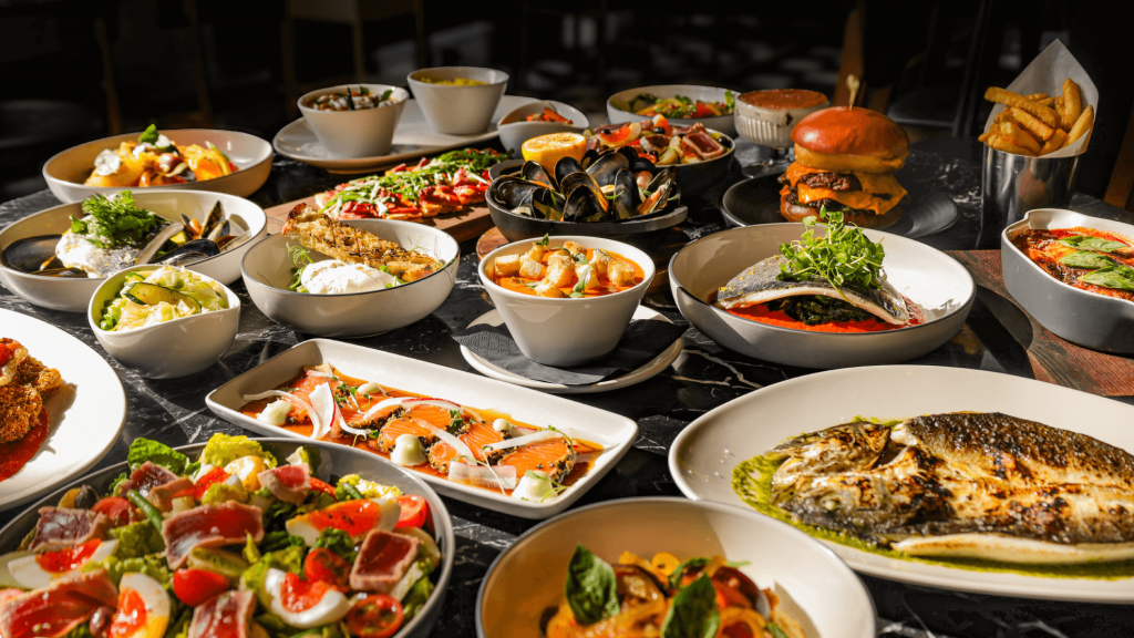 A table set out with a variety of plates of food from Bread Street Kitchens' spring menu. Seafood, salads, burgers, soups and sharing plates.