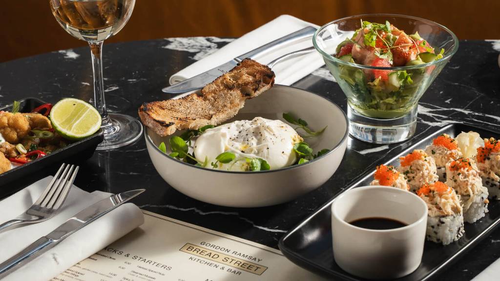 Bread Street Kitchen restaurant food spread featuring burrata, fresh salad, sushi rolls and white wine on black marble table with menu.