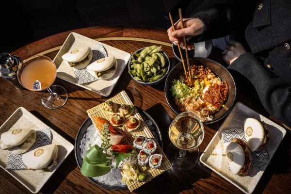 A person sat at a table eating a selection of Asian food, including bao buns, edamame, sushi, and noodles with chopsticks